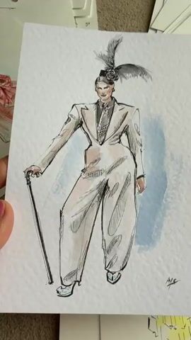 A hand holds up a watercolor sketch of a person wearing a light-colored suit, a hat with feathers, and holding a cane. Other sketches are visible in the background. The surface beneath the sketches appears to be carpeted.