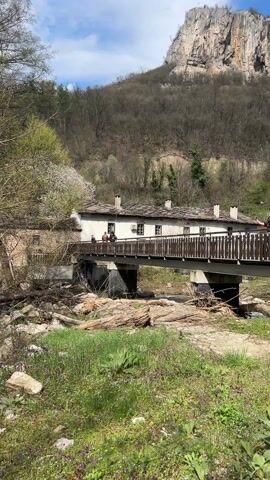 The video shows a scenic view of a small bridge over a river with a building next to it, set against a backdrop of a mountain. Several people are seen walking on the bridge, with a person in green standing out. The bridge has a wooden railing and supports. The building next to the bridge appears to be an old structure with a stone roof. The river below is partially blocked by logs and branches. The surrounding environment consists of trees and green grass. The sky is blue with a few clouds. The camera slowly pans to the right.