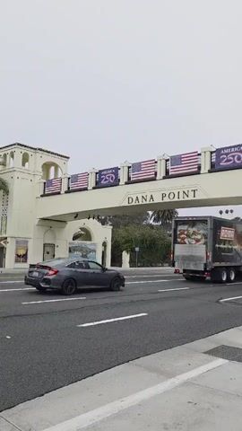 The video shows a street scene in Dana Point, California. An overpass with American flags and banners reading "AMERICA 250" spans the road. Cars and a delivery truck are driving on the road. A building with a tower is visible on the right side of the street, with a bus stop sign and advertisements in front of it. People are seen walking on the sidewalk. The sky is overcast.