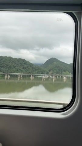 A view through a train window shows a wide, calm body of water reflecting a cloudy sky and a distant landscape. Across the water, a long bridge with multiple supports spans the expanse. Behind the bridge, lush green, tree-covered mountains rise, with a prominent peak in the center. The water's surface is smooth, creating clear reflections of the bridge and mountains. The sky is overcast with thick, gray clouds. The window frame, a light gray plastic, is visible in the foreground, framing the scenic view.