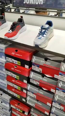 The video shows the interior of a shoe store, specifically a display of Skechers brand footwear. In the foreground, rows of Skechers shoe boxes are stacked on shelves. The boxes are predominantly white with blue and red Skechers branding. On the shelves above the boxes, several pairs of Skechers shoes are displayed.

At the beginning of the video, a pair of red and gray athletic shoes with white soles are prominently featured on a shelf. As the video progresses, the camera pans across different displays. We see various styles of Skechers shoes, including athletic sneakers for children, some with bright green accents and light-up features. The shoes are presented on white shelves. In the background, there are more shelves filled with shoes and shoe boxes, as well as a large display screen with the words "JUNIOR" visible.

There are a few people visible in the background, appearing to be customers browsing the store. One person, a woman with blonde hair, is seen from the back looking at shoes on a higher shelf. Another person, a man wearing a dark shirt, is also visible in the background, seemingly examining shoes. The lighting suggests an indoor retail environment. The overall impression is a typical retail setting focused on selling athletic footwear.