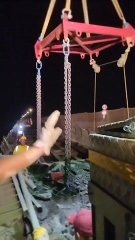 This video captures an industrial scene at night, likely a construction site. The focus is on a heavy-duty lifting apparatus consisting of a red frame suspended by straps and chains. Two individuals are actively engaged in guiding and positioning a large, heavy object, possibly a concrete segment or industrial component. One person, wearing a blue shirt and a white hard hat, is seen with their left arm extended, using a long pipe or pole to maneuver the object. They are wearing gloves. The second person, dressed in a bright yellow-green safety vest and a red cap, is crouched down and also appears to be assisting with the positioning, holding a pole or bar and wearing gloves. In the background, another person in a yellow safety vest can be seen further down the construction area. The environment is dimly lit by artificial lights, highlighting construction materials like pipes and rubble on the ground, and a long stretch of what appears to be railway tracks or a similar infrastructure project. The overall impression is one of controlled, heavy-duty work in a challenging, nighttime environment.