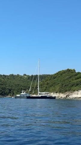 The video shows a calm, blue ocean under a clear sky. In the distance, there are two yachts anchored near a lush, green, tree-covered coastline. The larger yacht, a sailboat, is positioned in the foreground, and a smaller, white yacht is behind it. The coastline features rocky outcrops and vegetation. The camera is positioned on the water, looking towards the yachts and the shore. The water is gently rippling, indicating a peaceful environment.