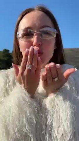 A woman with long, straight brown hair, wearing glasses, a white t-shirt with a silver cross necklace, and a white, fluffy, fringed jacket, presents her hands to the camera and blows kisses. She has square-tipped nails with a pale pink and white design and wears rings on her fingers. She turns her head to look to her left and then back to the camera. The background features a clear blue sky, a dark green hillside with trees, and a stone structure. Dance music plays in the background.