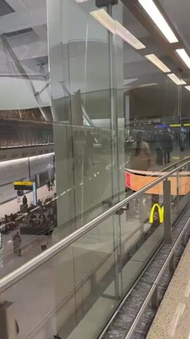 A first-person perspective video shows a walk through an airport terminal. The camera is moving forward at eye level. To the left, a glass railing overlooks a lower level of the terminal. On the lower level, several shops and restaurants are visible, including a McDonald's. People are walking around, and a few are sitting. To the right, a wide walkway with a marble floor stretches forward. A few people are walking on this walkway, and there are tall white columns supporting the ceiling. The ceiling is curved and has a series of lights. The overall environment is bright and modern. The video appears to be taken during the daytime.