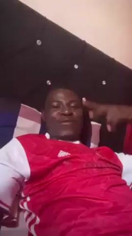 A man lies on a bed and speaks to the camera. He is wearing a red and white soccer jersey. The headboard is tufted and decorated with faux diamonds. He smiles and laughs at the camera. A curtain is slightly visible on the right side of the screen. The room is brightly lit.