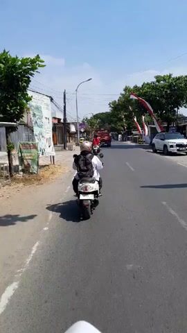 A motorcycle with two riders, one in front and one behind, travels down a paved road. The rider in front is wearing a white shirt and a black backpack, while the rider in the back has a black helmet. The motorcycle is moving away from the camera. The road is lined with buildings and trees on both sides. There are electrical wires overhead. On the left side of the road, there is a sign with text in Indonesian. On the right side, there are several flags with red and white stripes. A red truck is visible in the distance, also traveling on the road. The sky is blue with some clouds. The sun is shining, casting shadows on the road.