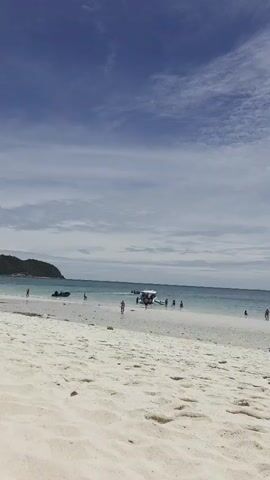 A vertical shot of a tropical beach under a partly cloudy sky. In the foreground, there is a sandy beach with footprints. In the midground, the turquoise ocean meets the white sand. Several people are visible walking along the beach and swimming in the water. Two boats are anchored in the ocean, one closer to shore and the other farther away. The overall scene depicts a peaceful day at a beach resort.