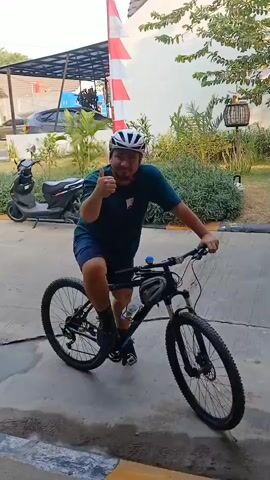 A man wearing a dark green t-shirt, blue shorts, a white helmet, and black shoes is riding a black mountain bike. He is on a paved surface, possibly a sidewalk or driveway, with a slight incline. In the background, there is a building with a white and red striped flag, a scooter, various plants, and some outdoor furniture. The man is looking to his right and appears to be pointing with his left hand. He has a water bottle attached to the frame of his bicycle and a small bag attached to the handlebars. The lighting suggests it is daytime, possibly late afternoon.