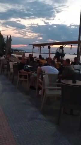The video shows a street scene near a body of water at dusk. People are seated at tables outside a cafe or restaurant. The sky is cloudy with hints of pink and orange from the setting sun. The ground is paved with tiles in a patterned design. In the foreground, a woman in a black dress and white shoes walks away from the camera. Further down the street, other people are walking and sitting. The atmosphere appears relaxed and social.