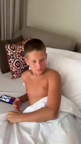 A young boy with a fair complexion and short brown hair sits shirtless in bed, covered with a white blanket, while someone off-screen, wearing a white shirt, applies a white cream to his face. The boy appears slightly uncomfortable as the cream is applied. Next to him on the bed is a smartphone displaying a video. The bed is covered in white linens, and behind it is a gray headboard and a few decorative pillows with geometric patterns. A cream curtain is visible in the background.