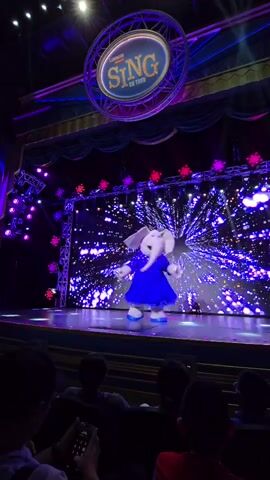 A short video clip shows a stage performance. A character, appearing to be a white elephant in a blue dress, is on stage. The elephant is facing away from the camera, and appears to be bowing or curtseying. The stage is lit with purple and blue lights, and the backdrop displays a dynamic, sparkly pattern resembling fireworks or a starburst. A circular sign above the stage reads "SING ON TOUR". Several audience members are visible in the foreground, some holding phones. The overall impression is a lively and colorful theatrical show.