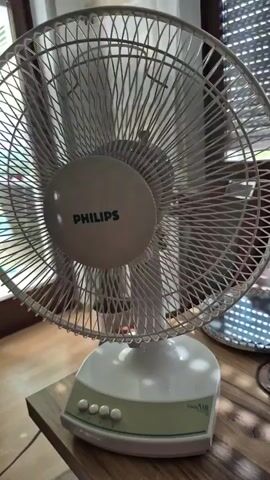 A close-up shot shows a white Philips brand fan with a green and white base sitting on a wooden table. The fan is oscillating, with its blades a blur as they spin. In the background, a large plant in a white pot is visible to the left, and blinds cover a window to the right. The scene appears to be indoors, likely in a room where the fan is being used to cool the air.
