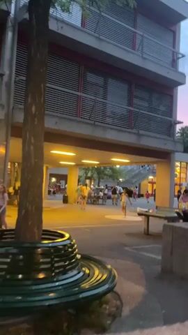 The video pans across an outdoor public space, possibly a plaza or courtyard, with a multi-story building as the backdrop. The building has a modern design with horizontal louvers on its upper floors. Below the building, there is a covered area lit by bright yellow lights, illuminating a gathering of people. In the foreground, a large, curved, dark green bench with a metallic pattern is visible, partially obscured by a tree trunk on the left. Further into the scene, people are casually interacting, some standing, some sitting at tables, and others walking. A ping-pong table is visible on the right side of the frame. The sky above displays a soft pink and blue twilight hue, indicating it is either dusk or dawn. The overall atmosphere appears relaxed and communal.