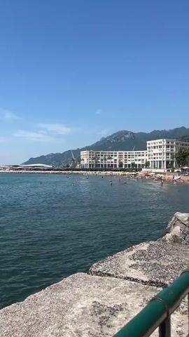 The video shows a body of water under a clear blue sky with scattered white clouds. In the distance, there is a coastline with buildings and mountains. Several boats are visible on the water, and a few people can be seen swimming or relaxing on the beach. The camera is positioned near the shore, looking out towards the sea.