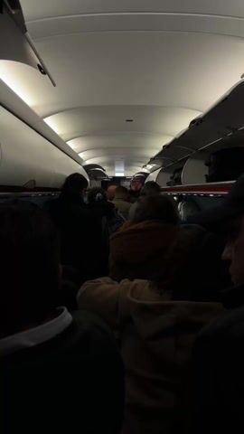 The video shows the interior of a plane cabin from the perspective of a passenger. The cabin is crowded, with people standing in the aisle, presumably waiting to deplane. Overhead luggage compartments are open, with some items still inside. The lighting is dim, creating a somewhat shadowy effect. Passengers are visible, mostly from the back, wearing various clothing. The general atmosphere appears to be one of anticipation as people prepare to disembark the plane.
