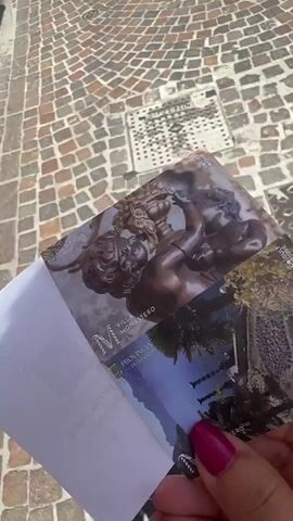 A hand with bright pink fingernail polish is holding two tickets with images on them. The tickets appear to be for Villa Monastero. The background is a cobblestone walkway. The image on the top ticket is a close-up of a bronze cherub sculpture. The image on the bottom ticket shows a pathway lined with tall, thin objects, possibly wrought iron fences or statues, with purple flowers in the background and a body of water and mountains in the distance. The tickets are partially obscured by a white piece of paper, which has some text and a QR code visible on it. The sun is shining, casting shadows on the ground.