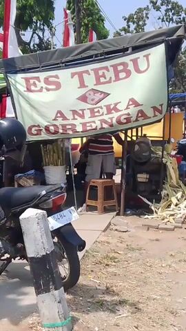 The video shows an outdoor food stall with a banner that reads "ES TEBU ANEKA GORENGAN". A person wearing a striped shirt and shorts stands behind the counter. To the left, there is a motorcycle parked next to a pile of sugarcane stalks. To the right, there are more sugarcane stalks and what appears to be fried food. The stall is decorated with Indonesian flags. The environment appears to be a street on a sunny day.