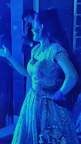 The video shows a woman in a traditional Indian outfit, a lehenga, dancing. She is wearing a sparkling top and a long skirt. She has a necklace and earrings. Her hair is tied up. There are people in the background, one of whom is a man. The lighting changes from blue to red to green and back to blue, creating a dynamic effect. The woman's movements suggest she is performing a dance. The environment appears to be a stage or a performance area.