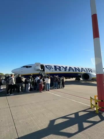 The video captures an outdoor scene at an airport tarmac on a clear, sunny day. The primary subjects are a large group of people and a white and blue Ryanair airplane.

In the first shot, a crowd of passengers is gathered around the front left side of the Ryanair aircraft, actively boarding or waiting to board via a mobile staircase. The plane's door is open, and some individuals are already ascending the stairs. The blue and yellow "RYANAIR" logo is prominently visible on the fuselage. The sky above is a brilliant, cloudless blue.

The second and third shots pan across the tarmac, showing a longer queue of passengers extending away from the airplane. They appear to be walking towards the boarding area or waiting in line. In the background, modern airport buildings with large windows and grey facades are visible under the clear sky. The ground is an asphalt or concrete tarmac with red and white markings. The overall impression is one of organized transit, with passengers preparing to depart on a flight from what is known to be Weeze, Germany. The time of day is daytime, evidenced by the bright sunlight and lack of artificial lighting. No strong emotions are discernible from the crowd; they appear to be typical travelers awaiting their turn.