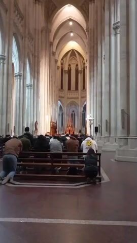 The video shows the interior of a large, ornate church. A group of people are kneeling on pews, facing the altar at the far end of the church. The architecture features high vaulted ceilings, arched windows, and tall stone columns. The lighting is dim, with some artificial light sources illuminating the space. The camera is positioned behind the kneeling individuals, focusing on the architectural details and the solemn atmosphere.
