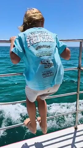 A person with blonde hair is hanging onto the railing of a boat, facing away from the camera. They are wearing a light blue t-shirt with graphics on the back, and light-colored shorts. Their feet are visible, and they appear to be barefoot. The boat is moving through turquoise water under a clear blue sky. The water is choppy, with white spray visible behind the boat. The railing is made of metal poles and appears to be a safety feature of the boat. The t-shirt graphics include text that appears to say "Monster Trucks," "Gunster," "Race Ace," and "Mega Wrex."