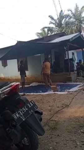 The video shows a rural scene with a house and a motorcycle in the foreground. Two men are standing on a blue tarp in front of the house, and a third man is standing behind them. There is a motorcycle parked on the left side of the frame. The house appears to be simple, with a tin roof and white walls. There are palm trees in the background. The overall atmosphere is casual and relaxed.