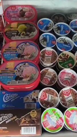 The video shows a close-up, eye-level view of an open freezer stocked with various brands and types of ice cream. The freezer is organized with rows of ice cream tubs and smaller individual servings. On the left side of the freezer, there are larger tubs of ice cream, including what appears to be Wall's brand products, with some labeled "Dessert Time" and one possibly showing a "3-in-1 Neo Trio" flavor. Below these, there is a box of Cornetto mini ice cream cones, which appears to contain 12 vanilla chocolate cones. Further down on the left, a box labeled "Cornetto mini disc" with a chocolate theme is visible. The right side of the freezer is filled with smaller, round containers of ice cream. These include products labeled "Oreo" and several others with a circular red and white logo, possibly from Wall's, featuring flavors like "Cokelat Vanilla" and "Cokelat Cookies". At the bottom right, there are colorful, smaller cups of ice cream, some with cartoon characters on them, suggesting they are likely aimed at children. The lighting is bright, illuminating the ice cream products clearly. There are no people visible in the frame. The overall environment suggests a retail setting, such as a convenience store or supermarket freezer section.