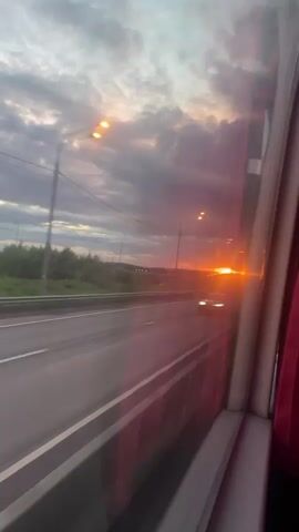 The video, shot from inside a moving vehicle, likely a bus due to the visible red curtain, shows a vivid sunset viewed through the window. The sky is a mix of dark, cloudy patches and lighter areas, with a bright orange sun sinking towards the horizon.  The vehicle is traveling along a highway with multiple lanes. Streetlights line the highway, and there is green vegetation and some structures in the distance.  A car passes in the same direction. The window is slightly tinted, giving the scene a reddish hue.