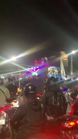 This is a vertical, low-light video taken at night, possibly at a street event or concert. The foreground shows the backs of several people on motorcycles, wearing helmets and dark clothing. The background features a stage with a DJ setup, bright colored lights (blue, green, pink, purple) flashing and moving. A large sound system is visible on stage. There are speakers stacked high on a structure. A crowd of people is gathered in front of the stage, and more people are on motorcycles. The overall atmosphere appears to be lively and energetic, with a focus on music and visual effects. The sky is dark, indicating nighttime. The scene is illuminated by the stage lights and headlights from the motorcycles.