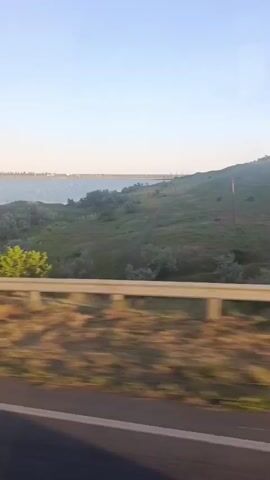 The video shows a view from a moving vehicle, likely a car or bus, looking out of a window to the right. The foreground consists of a blurred view of a road or shoulder with brown and green vegetation. A silver-colored guardrail runs horizontally across the lower portion of the frame. Beyond the guardrail, a grassy, undulating hillside dominates the mid-ground. The hillside is covered in various shades of green and brown vegetation, with sparse bushes scattered throughout. In the distance, a body of water, possibly a sea or a large lake, is visible under a clear, pale blue sky. The overall impression is of a scenic drive through a natural landscape.