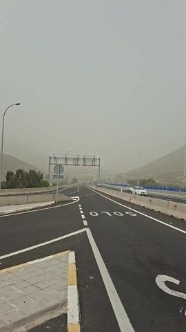 The video shows a road with a highway in the background. The sky is overcast with a brown haze. The road is black asphalt with white lines marking the lanes. There are road signs and barriers along the side of the road. Cars can be seen driving on the highway in the background.  There is a traffic direction sign in the middle of the road. A car makes a left turn at the end of the video.