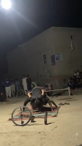 A group of people watch two men wrestle on a mat under a bright light at night. One wrestler is on his hands and knees, and the other is on top of him. There is a bicycle wheel next to the wrestlers.