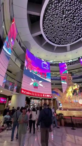 The video shows the interior of SkyCasino, a large, multi-story commercial space, with a high ceiling featuring a circular array of lights.  Multiple curved LED screens display video game-style graphics, seemingly showing gameplay. A gold statue of a horse with decorative elements is in the center of the space. People walk around the space, some looking at the displays and others passing through. The interior is well-lit with a clean, polished floor. Some people are interacting with their phones.