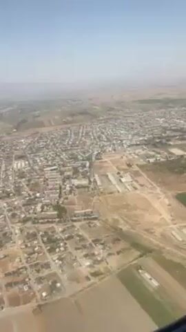 The video shows an aerial view of a town and surrounding landscape, taken from an airplane window. The town is characterized by closely packed buildings and a network of roads. Surrounding the town are fields, some green and cultivated, and others appearing drier and less vegetated. The sky is a clear, pale blue, suggesting a sunny day. The frame of the airplane window is visible in the lower right corner of the video, indicating the perspective from inside the aircraft.