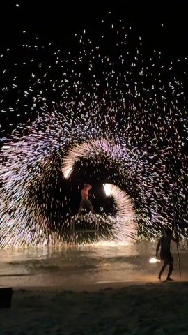 At night on a beach, a man stands in the shallow water, surrounded by a circle of sparks. Another man stands to the side, holding a torch. The performer spins the torch in a wide circle, creating a shower of sparks. The water reflects the light, and the sky is black.
