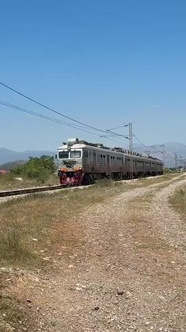 This media file features an electric train traveling along a railway track under a clear blue sky. The train is a passenger train, composed of multiple carriages, and appears to be moving from left to right across the frame. The locomotive at the front is light grey with red accents around the headlights and a prominent red and white graffiti tag across its front. The carriages are also predominantly light grey, with some sections showing wear and tear, and have red doors. Graffiti tags are visible on the sides of several carriages.

The scene is outdoors, with a natural setting. The railway track is laid on a gravel bed, flanked by dry, yellowish grass and scrub. In the background, rolling hills or mountains are visible, suggesting a somewhat rural or undeveloped area. The sky is a vibrant, cloudless blue, indicating a sunny day, likely late morning or early afternoon due to the bright illumination. The overall impression is of a functional, yet somewhat weathered, train journey in a sunny, arid landscape. There is no visible indication of people within the train or in the surrounding area. The location context provided is Mojanovići, Montenegro.