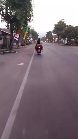 A first-person perspective video shows a person riding a motorcycle on a paved road. The rider is wearing dark clothing and is facing away from the camera. Another person is sitting behind the rider, also facing away. The motorcycle has a red taillight. The road is lined with trees and buildings on both sides. Colorful flags are hanging from poles along the road. The sky is overcast and the lighting suggests it is either early morning or late evening. There is a white line on the left side of the road. The motorcycle is moving forward on the right side of the road. The video is shaky, indicating movement.