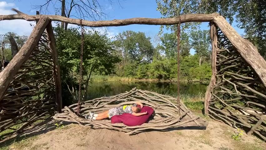 A person lies on a beanbag and a woven wooden swing in front of a body of water surrounded by trees. The swing has a rough, natural aesthetic, constructed from branches and logs. The person is wearing a t-shirt and shorts, with their head resting on a burgundy beanbag. The sun is shining, casting shadows, and the sky is clear and blue. The environment appears to be a park or a natural recreational area.