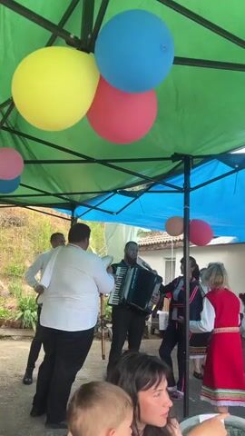 The video captures a lively scene of cultural celebration under a colorful tent. Three large balloons—yellow, blue, and red—hang from the tent's frame. A man in a white shirt stands holding a wind instrument, while another man in a black suit plays an accordion. People dressed in traditional Bulgarian clothing dance to the music. A woman in the foreground holds a young child, possibly her son, and appears to be watching the performance. The environment suggests a backyard or open space, and the overall atmosphere seems festive and joyful.