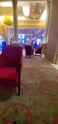 A person's legs and feet are visible in the foreground, clad in light-colored pants and dark shoes, resting next to a small, ornate table with a dark, spiral-shaped base and a marble top. The camera pans slightly, revealing several plush, red armchairs arranged in a seating area. In the background, a casino floor is visible with slot machines and patrons. People are seated at tables and walking around. The environment appears to be an indoor, upscale venue, possibly a hotel lobby or lounge area within a casino, characterized by ornate decor, including columns and chandeliers.