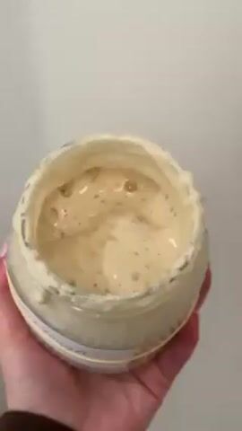A hand holding a glass jar against a white background is the focus of the video. The jar contains a beige-colored, bubbly substance. Some of the substance has spilled on the outside of the jar, near the top. The hand holding the jar has pink nail polish. The arm is wearing a brown sweater.
