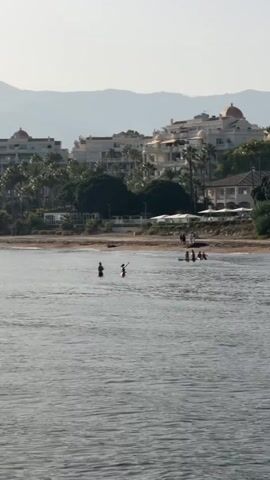 This is a landscape video filmed from the water, looking towards a beach and a town. The water is calm with ripples. In the foreground, there are several people wading or swimming in the water. In the middle ground, a sandy beach is visible with more people lounging and walking. Palm trees and buildings line the coast behind the beach. The buildings are a mix of white and pink, with some featuring red dome-like structures. In the background, mountains are visible. The overall scene depicts a sunny day at a coastal resort.