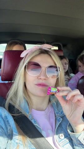 The video shows a woman in the front passenger seat of a car popping a piece of candy in her mouth. She has blonde hair, and is wearing a pink headband, sunglasses, a pink shirt, and a jean jacket. There are three other women in the car. One is sitting in the back behind the woman with the candy. She has blonde hair and is wearing sunglasses and a white t-shirt. Two young girls are in the back seat on the right side of the car. The car has dark red leather seats. The camera is held by the woman in the front seat. She makes eye contact with the camera and smiles.