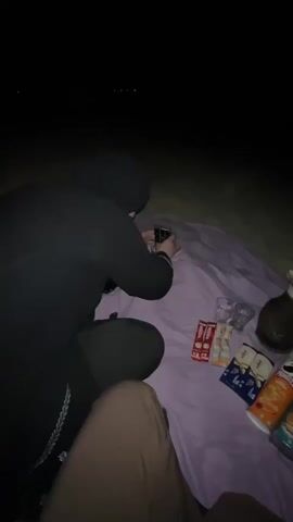 This video shows a nighttime scene on a beach. The camera is positioned at a low angle, looking up and to the right. In the foreground, a person wearing a black hijab and covering is kneeling on a purple blanket, holding a phone to take a picture or video. Beside them, on the blanket, are several items including Pringles cans, snack bars, and glasses. A thermos is also visible on the blanket. The background is dark, with some distant lights visible in the far distance, suggesting a shoreline and perhaps the ocean. The overall atmosphere is one of a quiet outdoor gathering or picnic in the dark.