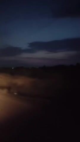 A low-light, slightly blurred video shows a dark, rural landscape at dusk or dawn. The sky is a deep blue with wispy, darker clouds. A faint orange glow, possibly from headlights or streetlights, illuminates a dusty or foggy area on the left side of the frame, creating indistinct shapes and streaks of light. The middle and right portions of the frame are predominantly dark, with the silhouette of trees or hills visible against the sky. There are no discernible people or specific objects in the foreground, and the overall impression is one of movement through a dimly lit, natural environment.