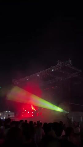 A crowd of people stands silhouetted against a stage at night. Red and green lights illuminate the stage with smoke. The stage is a complex metal structure with lights attached. The crowd is a dark mass of people, with some individuals standing out more than others. There is a hint of buildings in the far distance.