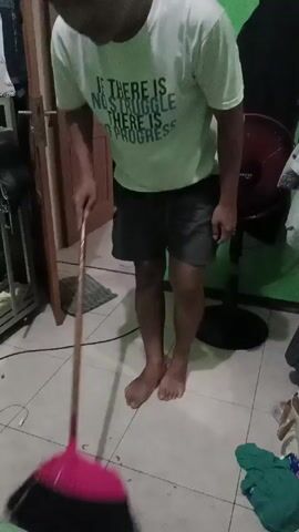 A barefoot person stands in a room with tiled floors, sweeping with a broom. The person wears a white T-shirt with a black graphic on it and dark shorts. The broom has a red handle and a black head. The room has a green wall, a brown door, and a black fan in the background. There are also some clothes, a metal shelf, and a white cable visible in the room. The person sweeps some spots off the floor.