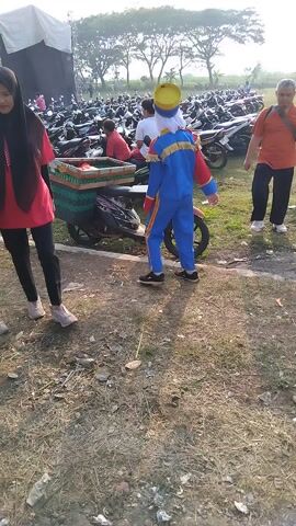 In a sunlit outdoor scene, likely in Sembojo, Indonesia, a group of people are gathered, potentially for an event. A young person in a vibrant blue and yellow marching band uniform, complete with a matching hat, stands near a red scooter. Their pose suggests they might be waiting to perform or are enjoying the festivities.

To their left, a person in a pink hijab and red top walks away from the camera, their posture indicating movement. On the right, an adult in an orange shirt and dark pants also moves, their face turned away. In the background, a large crowd is visible, with many motorbikes parked, suggesting a public gathering. A stage structure is also present, hinting at a performance or concert.

The ground is covered in dry grass and scattered debris. The sunlight casts shadows, indicating it is likely daytime, possibly late afternoon given the warm light. The overall atmosphere appears to be one of casual observation and community gathering, with the marching band uniform adding a touch of celebratory flair to the scene. No visible text is present in the media file.