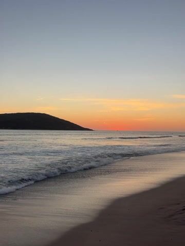 The video captures a tranquil sunset scene at a beach, likely in Mazatlán, Mexico. The foreground features foamy white ocean waves gently breaking and receding onto a smooth, sandy shore. The wet sand reflects the ambient light, creating a shimmering effect.

In the mid-ground, the vast expanse of the ocean stretches towards the horizon. A prominent dark headland or hill, covered in lush vegetation, is visible on the left side of the frame, extending into the water. The sky displays a beautiful gradient of colors, transitioning from a pale blue at the top to soft yellows and vibrant oranges closer to the horizon.

The setting sun, a distinct orange disc, is positioned low on the horizon, casting a warm, soft glow across the water and sky. A few small, dark figures can be seen in the distant water, possibly people swimming or surfing. The overall weather appears clear and calm, with minimal cloud cover. The scene evokes a sense of peace and natural beauty at the end of the day. No text is visible in the video.